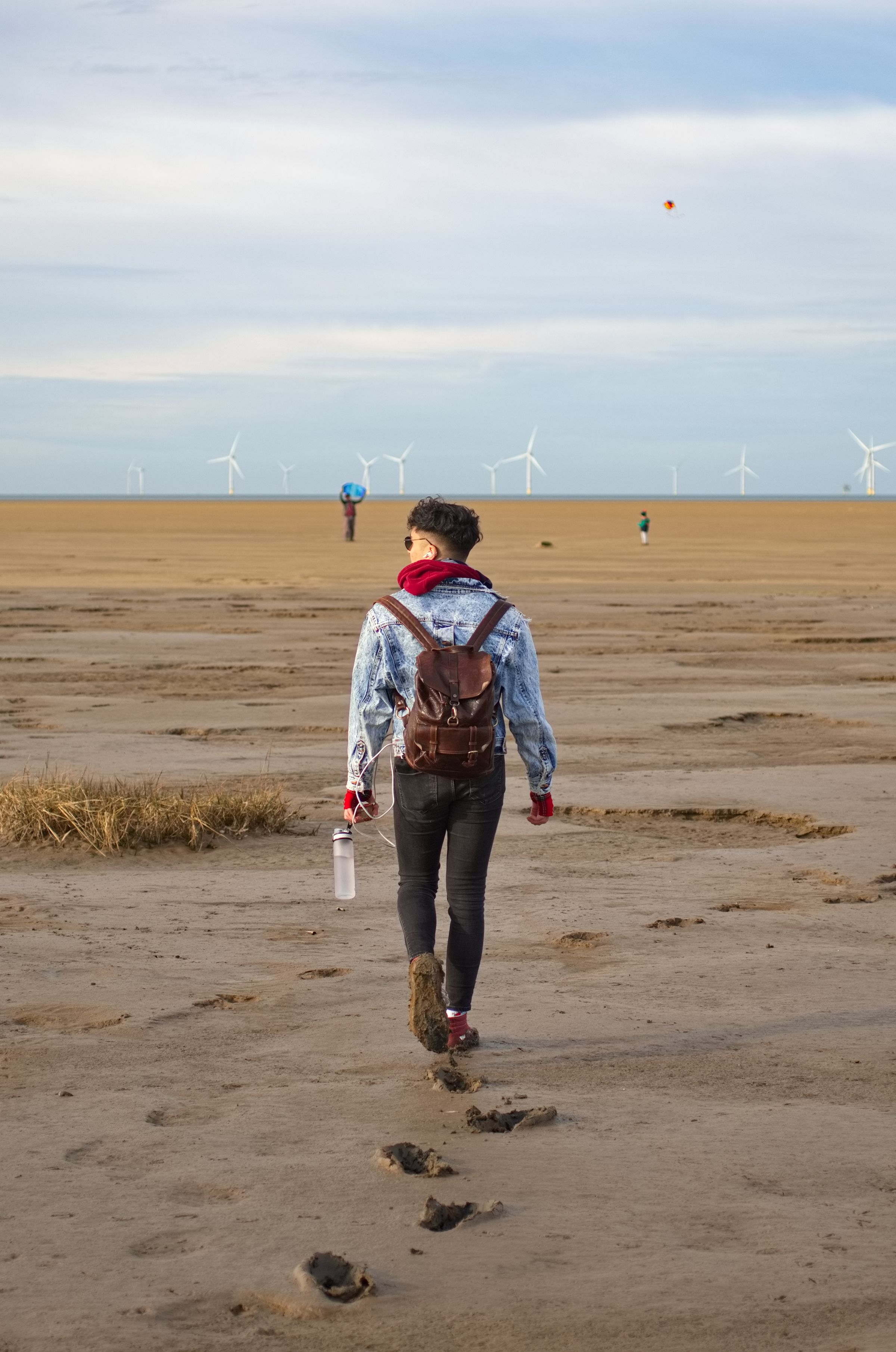 A man walking away from the camera towards a windfarm on the beach.