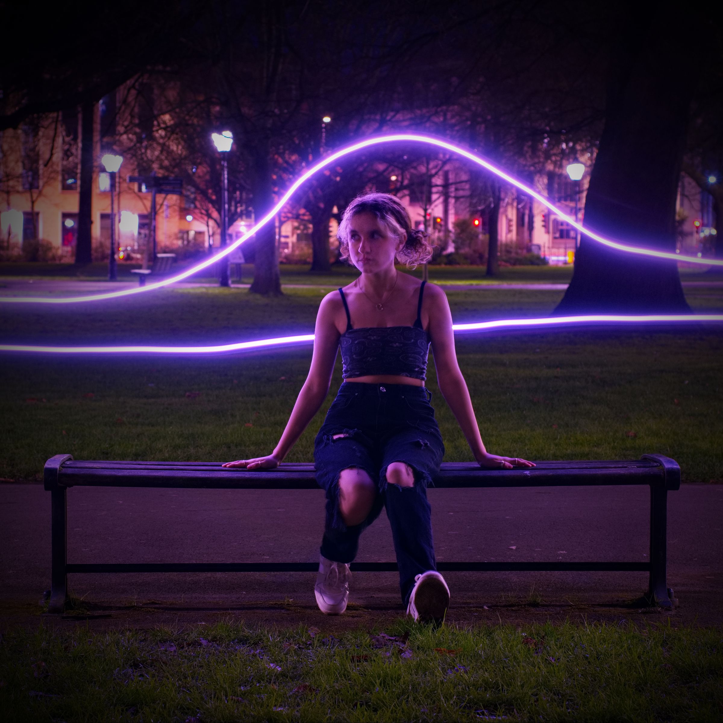 A woman sat on a bench in the dark, with two horizontal flourescent lines of light in the background.