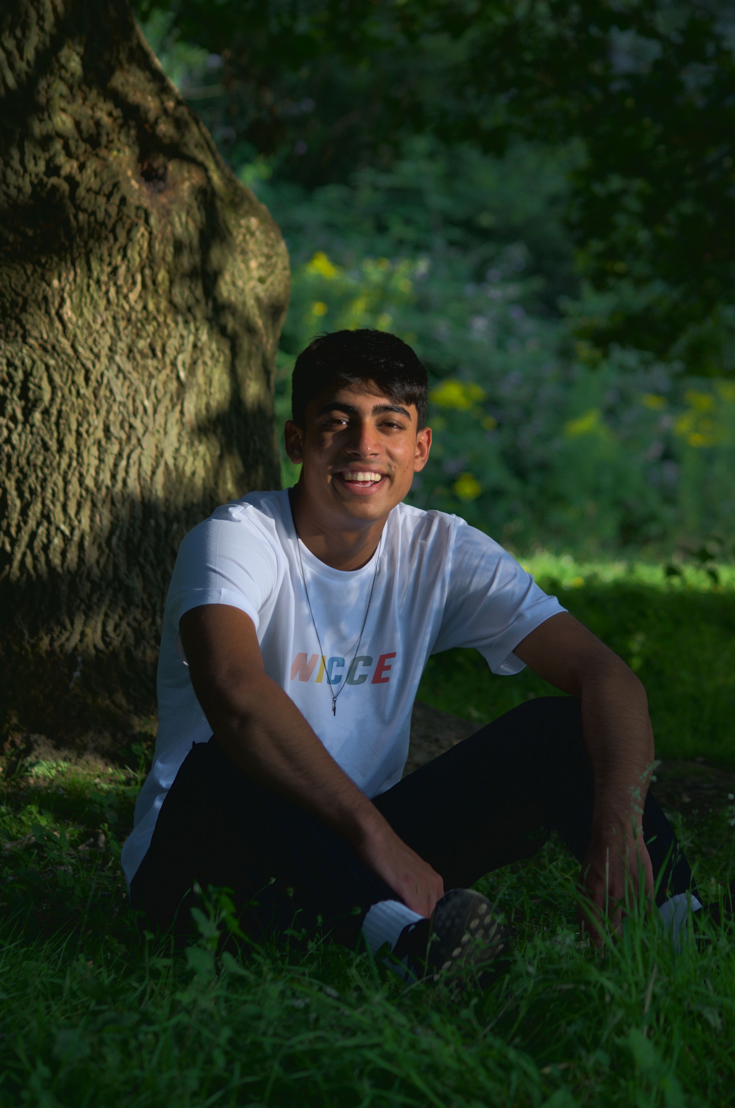 A man sat in dappled sunlight under a tree looking at the camera.