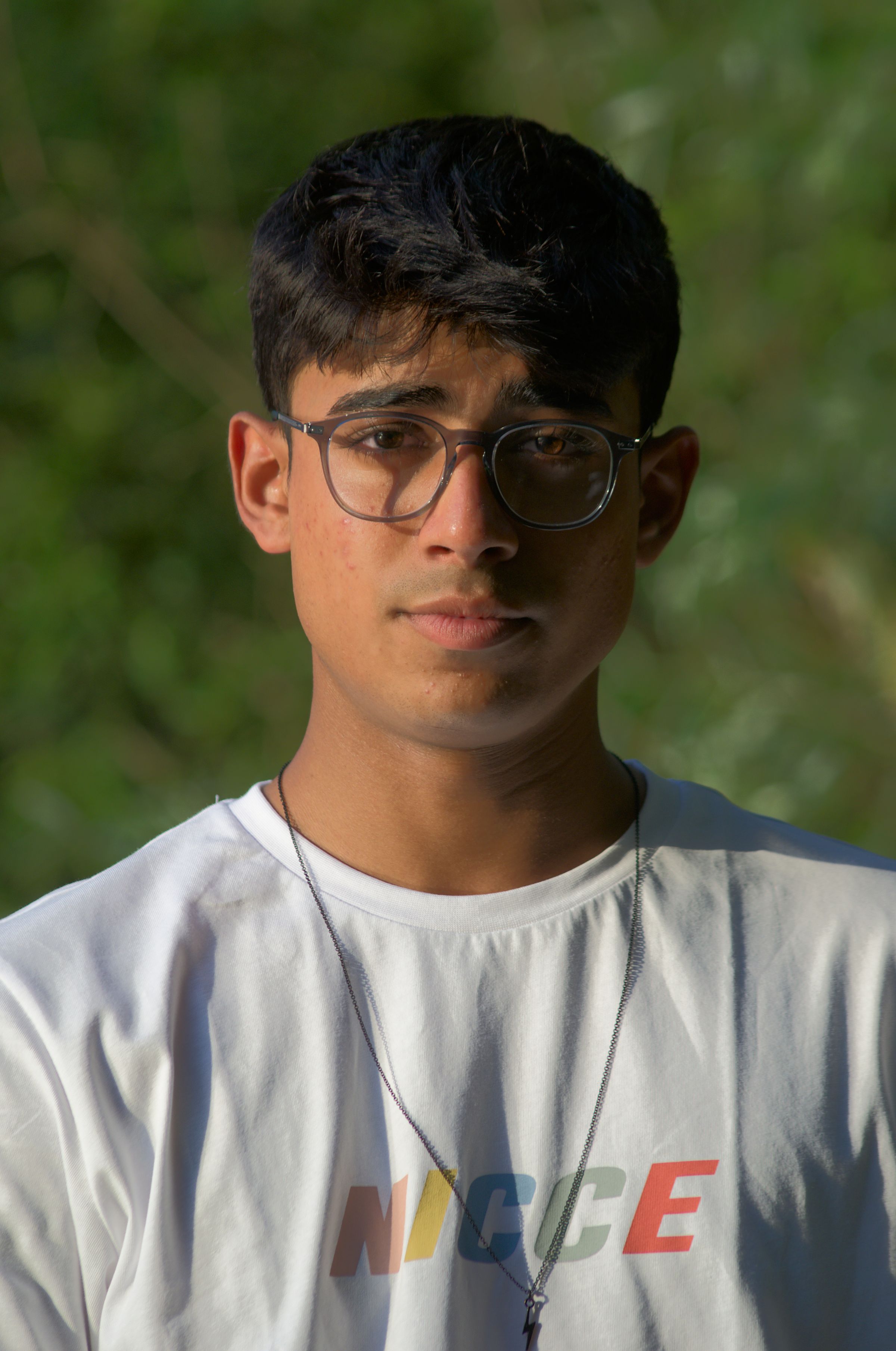 A man wearing glasses looking at the camera, with a leafy background.